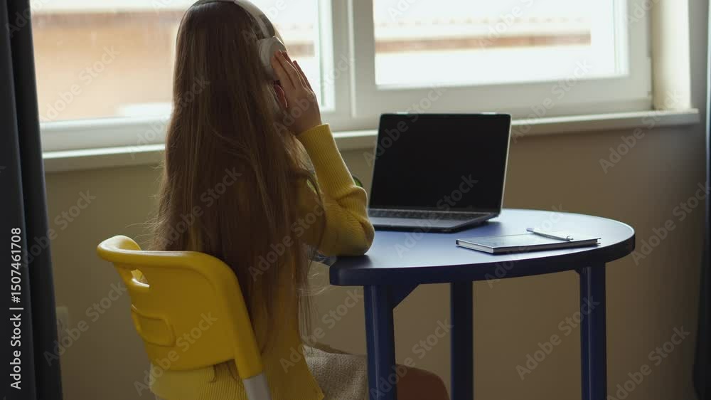 Poster rear shot of a person in a yellow sweater and headphones using a laptop on a blue table by a window.