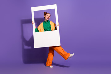 Cheerful young woman posing playfully with frame on vibrant purple background, showcasing trendy...