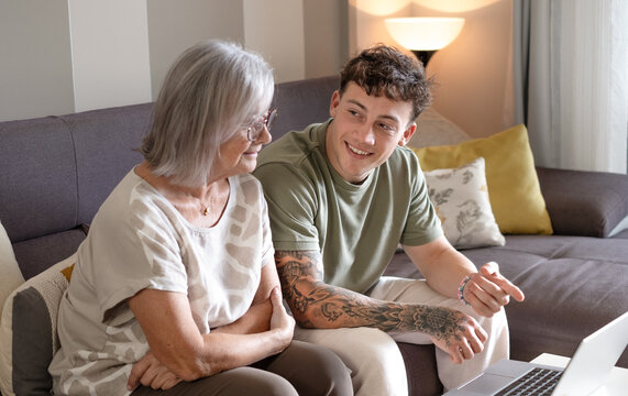 Happy young nephew teaching technology to his old grandmother using laptop. Senior woman learns to use computer, shopping online, make reservations. Multi generation family sitting on sofa