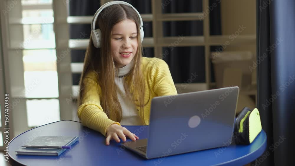 Poster a young girl sits at a round blue table, wearing headphones and using a laptop. she is smiling and a