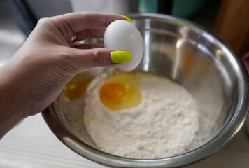 woman's hand holding an egg with the fingers over a metal mixing bowl filled with flour