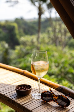 Cocktail with ice in a wine glass served with a dish of peanuts next to tortoise shell sunglasses with view of rice paddies in an open-air structure in Bali, Indonesia. 
