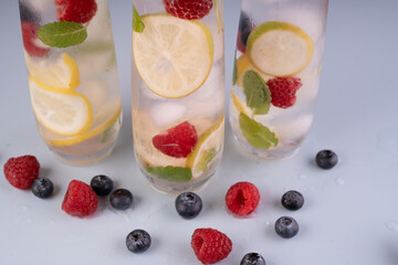 refreshing beverage display with lemon and berry infused water in tall glasses with mint leaves, surrounded by fresh blueberries and raspberries on a light blue surface, relaxing, pleasant, tranquil