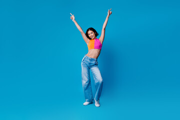 Energetic young woman striking a cheerful pose wearing stylish casual outfit against a bright blue...