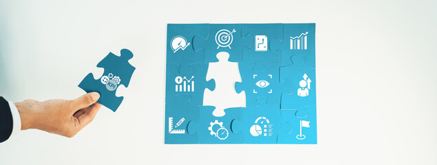 A hand holding a puzzle piece above a completed puzzle featuring various business icons, symbolizing strategies, analytics, and teamwork to achieve organizational goals. Amity