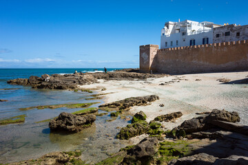 View of the historic medina wall in Essaouira, Morocco, by the rocky Atlantic shore at low tide. White buildings rise above the wall under a bright blue sky