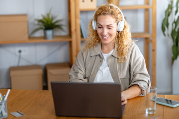 Smiling woman wearing headphones works on her laptop in a cozy home office setting, staying focused and engaged during an online meeting or remote work session.