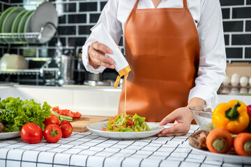 Asian housewife holding dressing bottle to pouring into fresh vegetable while wearing apron and standing