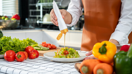 Asian housewife holding dressing bottle to pouring into fresh vegetable while wearing apron and standing