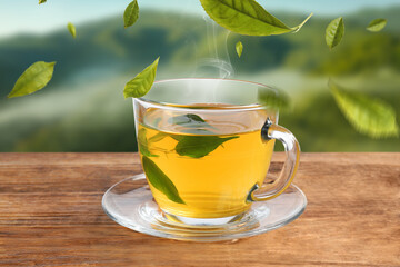 Green tea leaves flying above glass cup with hot beverage on wooden table in mountains