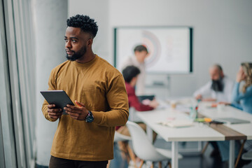 Pensive businessman holding tablet, observing meeting in modern office