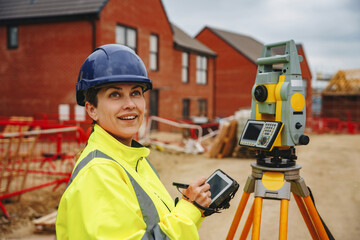 Woman engineer in  bright safety jacket and hard hat uses surveying instrument at a construction...