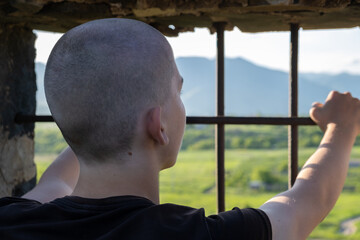 Rear view of a shaved-headed teenage boy in a solitary prison cell, leaning on the barred window and gazing at the sunset and mountains beyond. Longing for freedom and missing home.