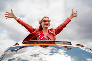 Woman with sunglasses joyfully enjoying a car ride with arms raised under a cloudy sky in a vibrant...