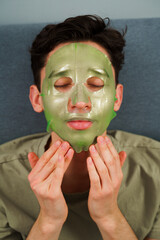 Relaxed man with hydrating face mask at home. Man is applying facial mask for rejuvenate...