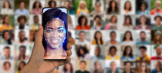 A person holds a smartphone displaying a face recognition scan in front of a collage of diverse faces. This showcases modern identification technology in action, highlighting its potential uses.