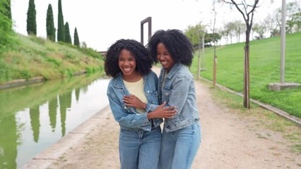 Two happy twin sisters wearing denim jackets and jeans, holding hands and walking near a canal in a city park - Powered by Adobe