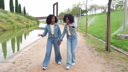 Two happy twin sisters wearing denim jackets and jeans, holding hands and walking along a canal in a green park - Powered by Adobe