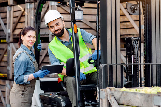 Distribution, loading of fruits for sale in store and market, work in warehouse during harvest season. Smiling young female show tablet to man in hard hat in forklift work, on wooden boxes background