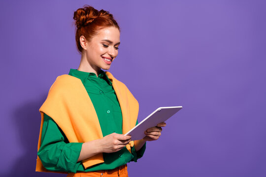 Lovely woman with red hair using a tablet against a purple background, wearing vibrant casual clothes and smiling effortlessly