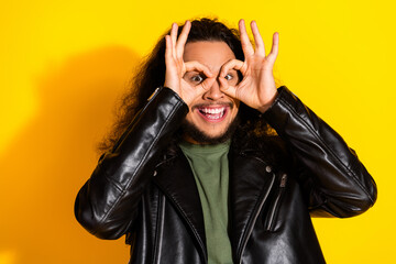 Cheerful young man in leather jacket making playful hand gestures on vibrant yellow background