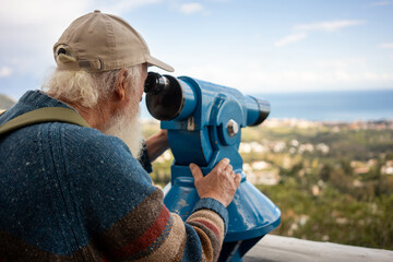 Close-up of senior man with white beard and cap using a public blue telescope to observe the scenic...