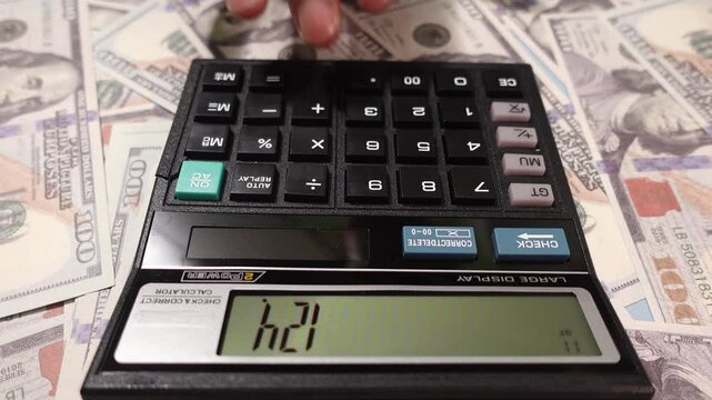 A hand operates a calculator surrounded by scattered US dollar bills.  Woman hands pressing calculator buttons analyzing business income.