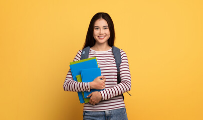 A cheerful Asian student stands confidently, clutching several colorful textbooks. She wears a striped shirt and has a backpack, exuding a sense of readiness for school and learning.