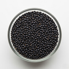 Close-up top-view of whole black urad dal (black gram) in a borosilicate glass bowl, scattered naturally, on white background