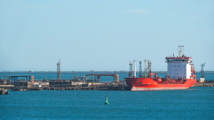 Large red tanker ship docked at an industrial port with pipelines and structures
