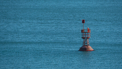 A red navigational buoy stands alone in calm blue ocean waters under natural light, marking a sea channel