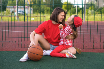 Funny laughing mom and daughter relax on the playground after playing basketball