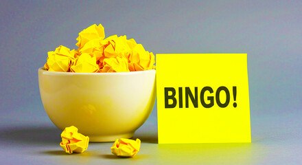 A bowl of yellow crumpled paper next to a yellow note with the word bingo written on it against gray