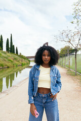 Stylish young woman with afro hair holding a smartphone, posing confidently in a vibrant park by a flowing river, enjoying the outdoors