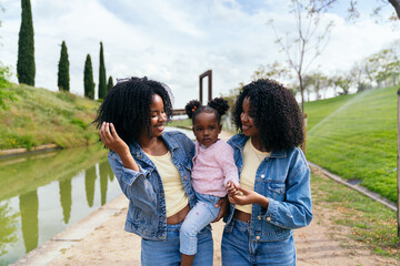 African twin sisters walking together in a city park, joyfully holding their niece while enjoying a...
