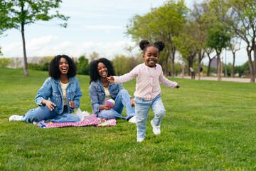 Cheerful toddler running on grass while her mother and aunt are sitting on blanket having picnic in...