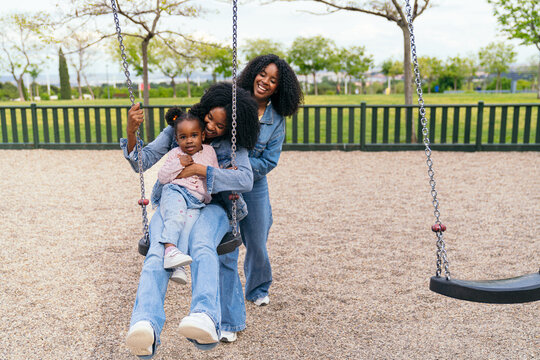 Mother and aunt joyfully pushing their niece on a swing in a vibrant playground, creating cherished memories on a sunny day