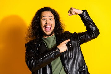 Charming young man wearing a leather jacket, pointing confidently to a flexed arm against a vibrant yellow background