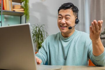 Asian man wearing headset, freelance worker, call center or support service operator helpline, having talk with client or colleague communication support at home room. Guy sits on couch at table desk