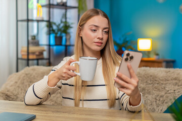 Caucasian woman sits at home at table, scrolling social media content videos on smartphone, sipping...