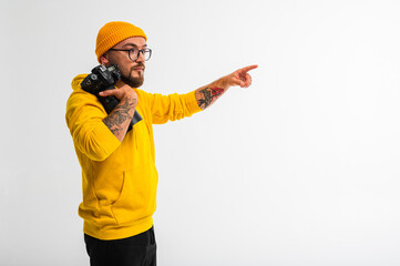 Stylish male photographer in yellow hoodie and cap, facing forward with camera in hand, pointing to the side with hand, place for inscription. Creative concept, studio portrait on white background.