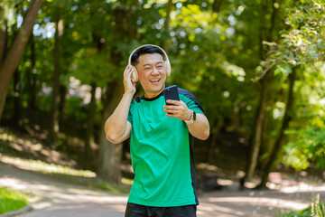 Athletic fitness adult guy sport runner man walking, wearing headphones listening music from smartphone and smiling. Healthy Japanese sportsman workout cardio outdoors in morning park on summer day.