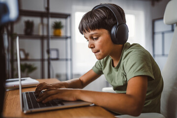 caucasian boy use headphones and laptop computer at home for online learning study or work on...