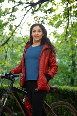 Happy smiling woman long brown hair biking on bicycle on spring trees nature in red jacket and...