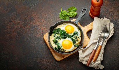 Two fried eggs with spinach in a cast iron pan on a wooden board with fresh ingredients, healthy...