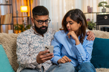 Indian couple man and woman sitting on home sofa, using smartphone for food order, joking and...