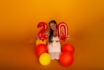 Young birthday girl wearing white outfit holding age number next to air balloons sitting against dark orange color background.