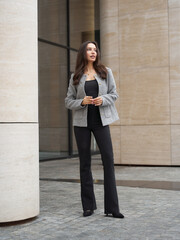 Obraz premium A woman with long hair stands outdoors in front of a modern building. She wears a gray blazer over a black top and black pants, holding a cup in one hand. Her expression is thoughtful