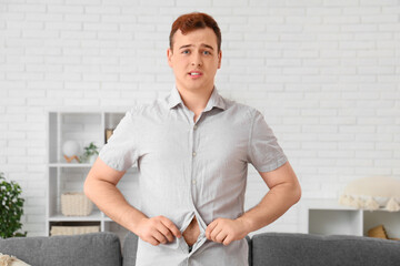 Young worried overweight man putting on tight clothes at home. Weight gain concept