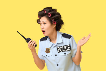 Female Asian police officer with hair curlers and radio transmitter on yellow background